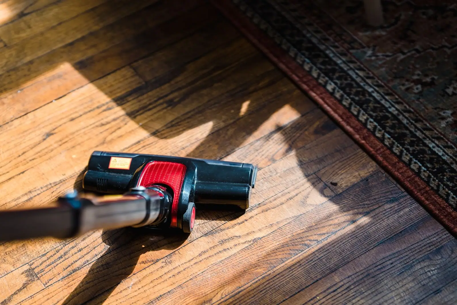Cordless vacuum cleaner on a wooden floor beside a patterned rug in sunlight